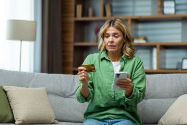 Kandırılmış üzgün kadın sahte ödemeler yapmaya çalışıyor oturma odasındaki kanepede oturuyor depresif banka kartı ve akıllı telefon tutarak.
