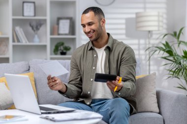 Evden bilgisayarla uzaktan çalışan gülümseyen İspanyol genç adam. İçinde belgeler, raporlar, hesaplar ve hesap makinesi var. Mali sonuçtan memnun..