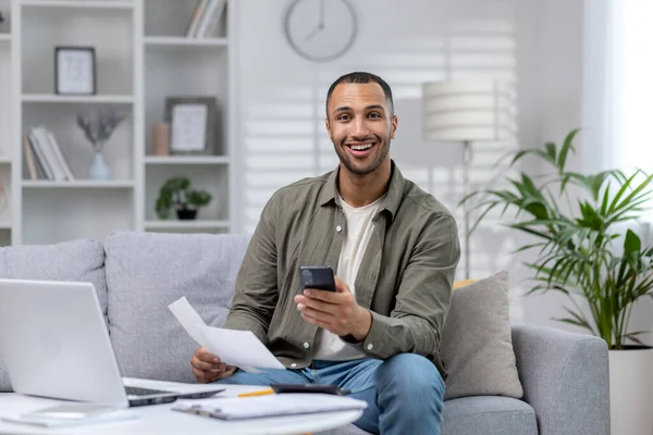 Portrait of a young African-American businessman who works remotely from home. He is sitting on the sofa in front of the laptop, holding a phone and documents in his hands. Smiling at the camera.