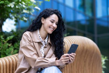 Cheerful beautiful woman sitting on bench outside office building, successful Latin American business woman using phone, reading news online and browsing online pages, holding smartphone.