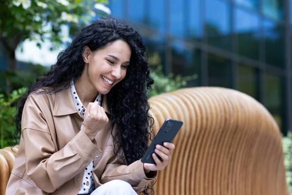Young happy woman celebrating victory triumph success, satisfied hispanic business woman holding phone received online win notification, sitting on bench outside office building.