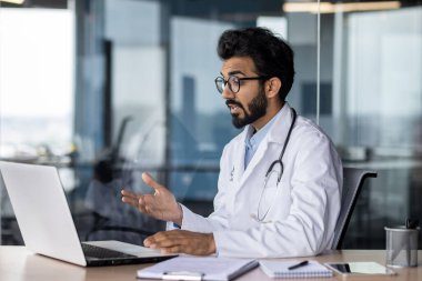 A young Indian doctor is sitting in the office at the table with a laptop and talking and consulting on a video call with patients. Holds an online meeting with colleagues.