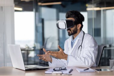Portrait of a young Indian doctor sitting at a table in a hospital, wearing a virtual mask.