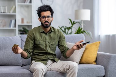 Tatminsiz ve hayal kırıklığına uğramış Hintli adam dolandırıcılar tarafından çevrimiçi aldatıldı, kameraya baktı, oturma odasında kanepede oturan adam banka kartı ve telefon tutuyordu..