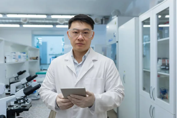 Portrait of a young Asian male scientist standing in a laboratory wearing protective glasses and gloves. He was holding a folder with documents in his hands. He looks seriously into the camera.