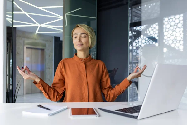 stock image Businesswoman resting at workplace meditating in lotus position sitting at the table inside the office, young successful female worker satisfied with the results of the of new projects and victories.