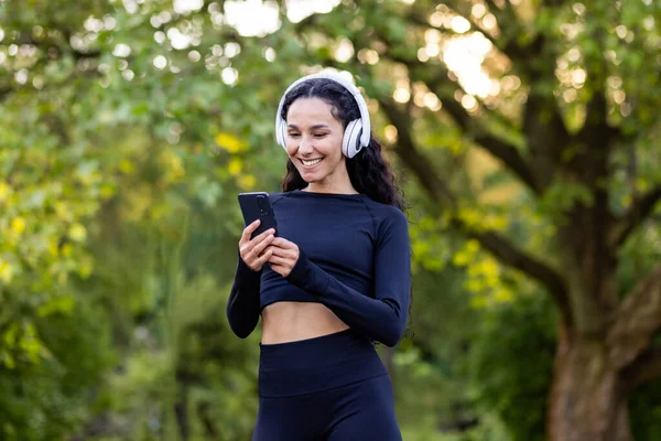 Young beautiful Latin American woman walks in the park during active physical exercises, woman uses an application for listening to audio books and music on phone, smiling sportswoman in headphones.