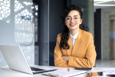 İş yerindeki olgun kadın patronun portresi, kollarını kavuşturup masada oturan iş kadını gülümseyen ve kameraya bakan, belgeli ve evraklı başarılı finansör..