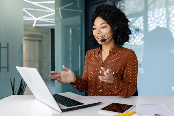 Young beautiful woman talking to customers remotely, african american female worker smiling using headset phone and laptop for online consultation, customer service.