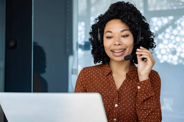 Young beautiful woman talking to customers remotely, african american female worker smiling using headset phone and laptop for online consultation, customer service.