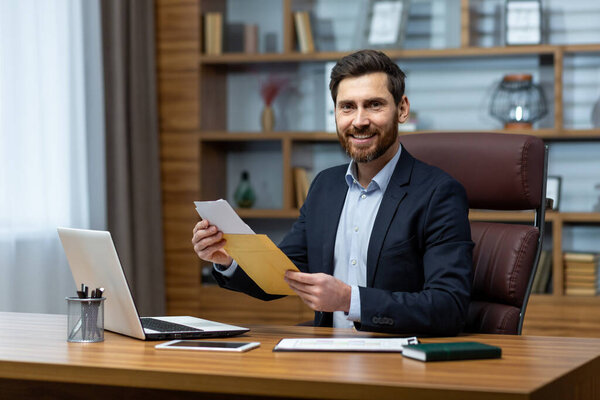 Портрет счастливого инвестора-бизнесмена, зрелого взрослого мужчины, улыбающегося и смотрящего в камеру, босса, держащего конверт с хорошими новостями внутри офиса на рабочем месте.