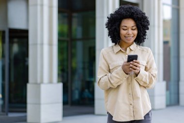 Telefonla gülümseyen genç Afrikalı Amerikalı kadın, ofis binasının dışındaki iş kadını akıllı telefon uygulaması kullanıyor, mesajlar yazıyor ve internet sayfalarını tarıyor..