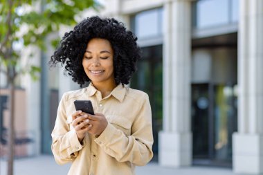 Telefonla gülümseyen genç Afrikalı Amerikalı kadın, ofis binasının dışındaki iş kadını akıllı telefon uygulaması kullanıyor, mesajlar yazıyor ve internet sayfalarını tarıyor..