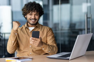 Başarılı İspanyol genç işadamının portresi, iş yerinde gülümseyen ve kameraya bakan bir adam zafer başarısını kutlayan bir telefon tutuyor, başarı başarısından memnun.