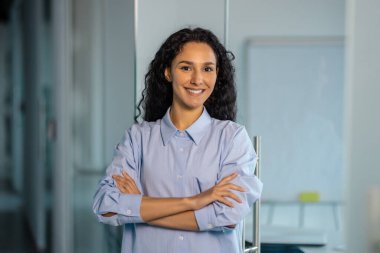 Mutlu ve başarılı iş kadını portresi, tişörtlü patron kollarını kavuşturup ofisin içinde kameraya bakıyor koridorda kıvırcık saçlı İspanyol kadın..