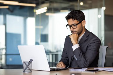 İş yerindeki genç ve düşünceli iş adamı, ofis içi dizüstü bilgisayarlı, gözlüklü ve takım elbiseli patron teknik finansal çözümleri düşünüyor, çenesinde dinleniyor..