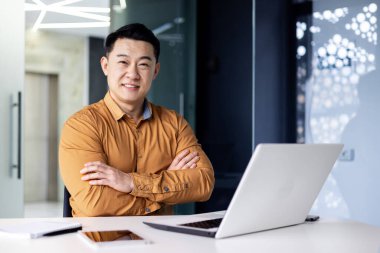 İş yerindeki deneyimli Asyalı iş adamının portresi. Patron programcı masada oturuyor. Kollarını kavuşturup gülümsüyor ve dizüstü bilgisayarla kameraya bakıyor..
