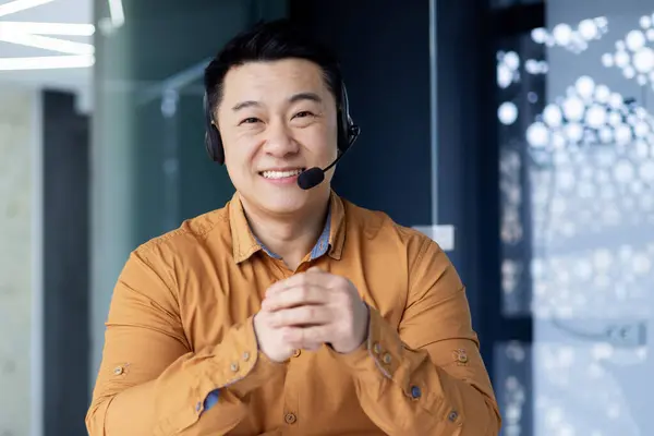 Portrait of a businessman customer support worker for online buyers, man at workplace smiling and looking at camera, asian man with headset for video call.