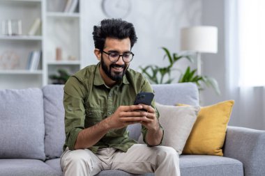 Genç gülümseyen adam evde oturma odasındaki kanepede oturmaktan mutlu, Hintli adam telefonda bir uygulama kullanıyor, bir mesaj çeviriyor, aramalar yapıyor ve internet sitelerini tarıyor..