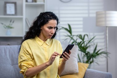 Üzgün ve hayal kırıklığına uğramış ciddi düşünceli bir kadın evde yalnız başına telefondaki uygulamayı kullanıyor. Yakın plan, İspanyol asıllı biri evdeki oturma odasında oturuyor..