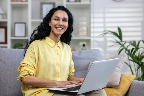 stock image Portrait of young beautiful hispanic woman at home with laptop, woman smiling and looking at camera sitting on sofa in living room at home.