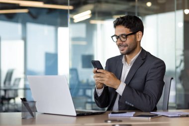 Başarılı deneyimli iş adamı iş yerinde otururken telefon kullanıyor, İspanyol asıllı gülüyor, başarıdan memnun, telefon tutuyor, internet üzerinden akıllı telefon uygulaması kullanıyor..