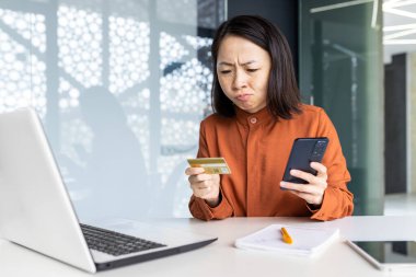 İş yerindeki üzgün ve hayal kırıklığına uğramış kadın, elinde banka kartı ve telefon tutan Asyalı kadın, iş kadını reddedildi ve para transferi hatası oldu..