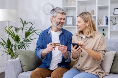 Olgun bir çift, evdeki oturma odasında birlikte oturuyor, neşeli bir aile online alışveriş ürünlerini seçiyor, online alışveriş için telefon kullanıyor ve banka kredi kartı kullanıyor..