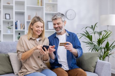 Olgun bir çift, evdeki oturma odasında birlikte oturuyor, neşeli bir aile online alışveriş ürünlerini seçiyor, online alışveriş için telefon kullanıyor ve banka kredi kartı kullanıyor..