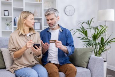 Üzgün ve aldatan çift, erkek ve kadın oturma odasında birlikte otururken hayal kırıklığına uğradı, telefonda online alışveriş uygulaması kullanarak, banka kredi kartını tutarak, para transferini reddederek,.