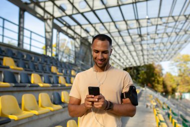 Erkek sporcu stadyumdaki beden eğitimi dersinden sonra telefonda uygulama kullanıyor, kulaklıklı antrenör müzik dinlemek, yürümek, gülümsemek ve dinlenmek için akıllı telefon kullanıyor..