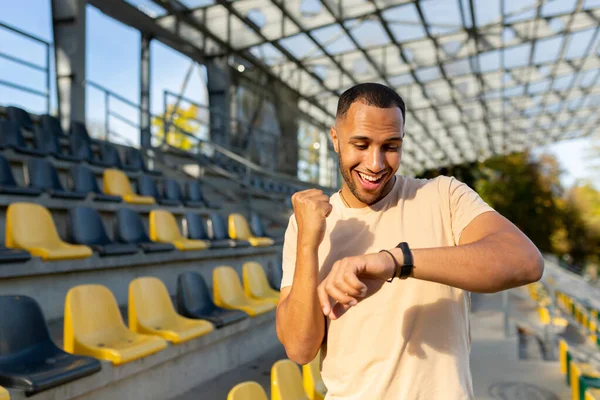 Happy and successful man satisfied with the results of physical training celebrating good results of fitness and running looking at smart watch fitness bracelet walking in the air in the stadium.