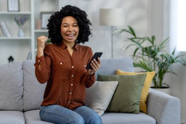 Mutlu Afro-Amerikalı kadın elinde telefonla koltukta oturmuş kameraya bakıp zafer jestini eliyle gösteriyor..