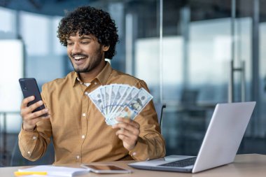 İş yerindeki işadamı mali başarı elde etmekten mutlu, elinde telefon ve dolar banknotları tutan, ofisin içinde dizüstü bilgisayarla çalışan bir adam..