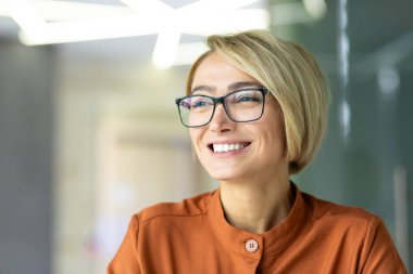 Hayalperest kadının yakın plan portresi, iş kadını gülümseyen ve iş yerindeki pencereden bakan, gözlüklü ve gömlekli sarışın..