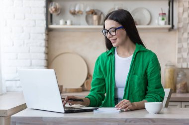 Genç bir kadın evde dizüstü bilgisayar kullanarak çalışır ve çalışır. Mutfakta, masada oturup bir deftere yazı yazmak..