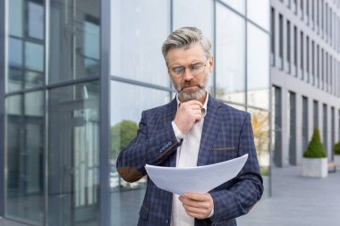 Ciddi düşünen işadamı patronunun belge sözleşmeleri ofis binası dışında faturalar, iş elbisesi giymiş olgun bir adamın evraklara bakması, ve mali raporlar..