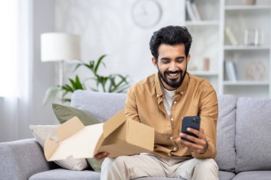Kanepede oturan neşeli adam bir paket aldı, Latin Amerikalı online mağazadan satın almaktan memnun, telefondaki uygulamayı kullanarak olumlu bir eleştiri yazdı.
