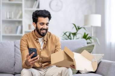 Kanepede oturan neşeli adam bir paket aldı, Latin Amerikalı online mağazadan satın almaktan memnun, telefondaki uygulamayı kullanarak olumlu bir eleştiri yazdı.