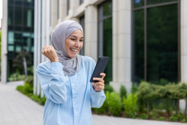 Şehre inen tesettürlü neşeli kadın, online galibiyet ve telefonda iyi haberler aldı, ofis binasının dışındaki iş kadını, zafer kazanan el kaldırdı..