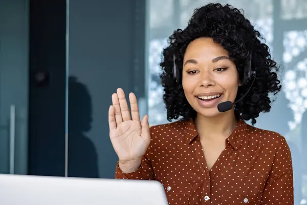 Woman with headset phone using laptop for remote communication and video call, african american woman waving at camera greeting.