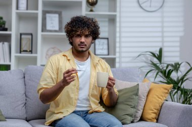 Soğuk ve ateşle evde hasta bir adam, oturma odasındaki koltukta oturan İspanyol, elinde ilaç ve vücut termometresiyle sıcak bir içecek tutuyor..