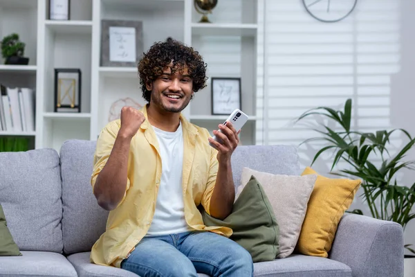 Portrait of young successful man at home on sofa, winner smiling and looking at camera holding phone in hands using game app, holding hand up successful gesture of triumph.