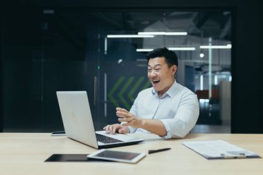 Asyalı genç işadamı, ofiste dizüstü bilgisayar ve belgelerle çalışıyor. Hevesle ve gülümseyerek video aracılığıyla iletişim kurar, başarılı bir anlaşma yapar, projeyi bitirir..