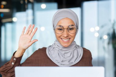 Genç tesettürlü, tesettürlü müslüman bir kadın bilgisayarla video üzerinden konuşuyor. Elini kameraya sallıyor, merhaba diyor..