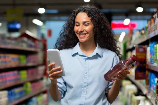 Portrait of a female buyer in a supermarket, a Hispanic woman chooses shampoo for hair and shower gel, reads reviews on the Internet, smiles and uses an application on her phone.