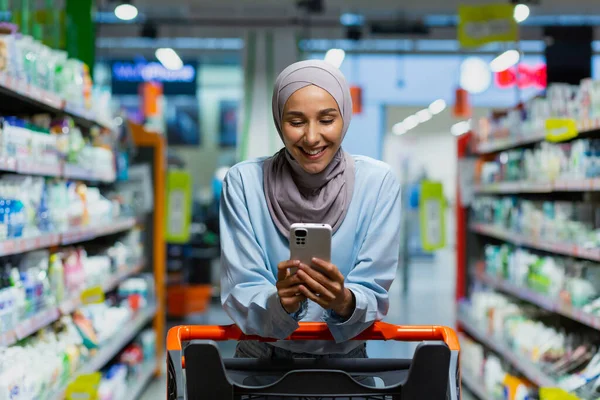 Portrait of a shopper inside a large supermarket store, a Muslim woman in a hijab with a shopping cart uses a smartphone app, chooses products with discounts and smiles.