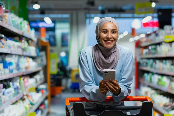 Portrait of a shopper inside a large supermarket store, a Muslim woman in a hijab with a shopping cart uses a smartphone app, chooses products with discounts and smiles.