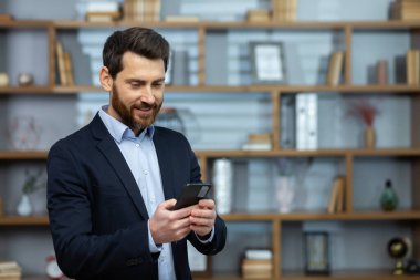 Ofisin penceresinin yanında duran başarılı, deneyimli iş adamı, telefon kullanan, mesaj yazan ve online sayfaları karıştıran patron, işadamından memnun..