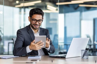 İşyerindeki genç başarılı İspanyol işadamı tablet bilgisayarda uygulama kullanıyor ofis binasında gülümseyen adam dizüstü bilgisayarla çalışıyor, iş elbisesi giyiyor, başarısından memnun..